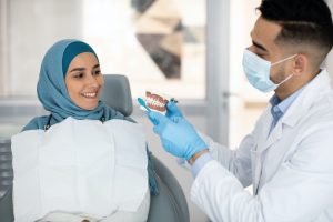 Male dentist holding jaw model, explaining dental hygiene to muslim female patient